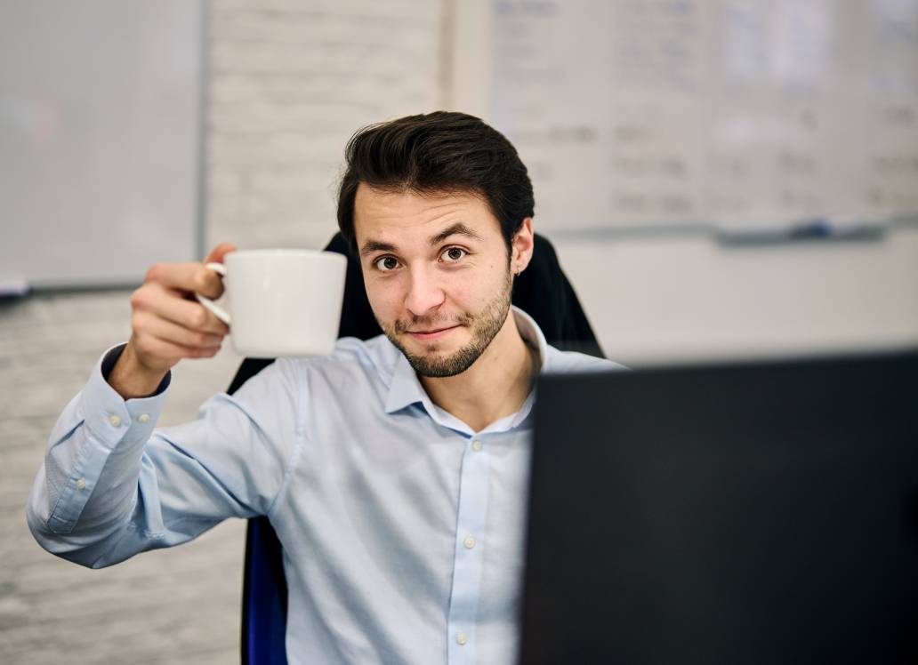 Man smiling and raising a coffee mug at his desk in an office setting.